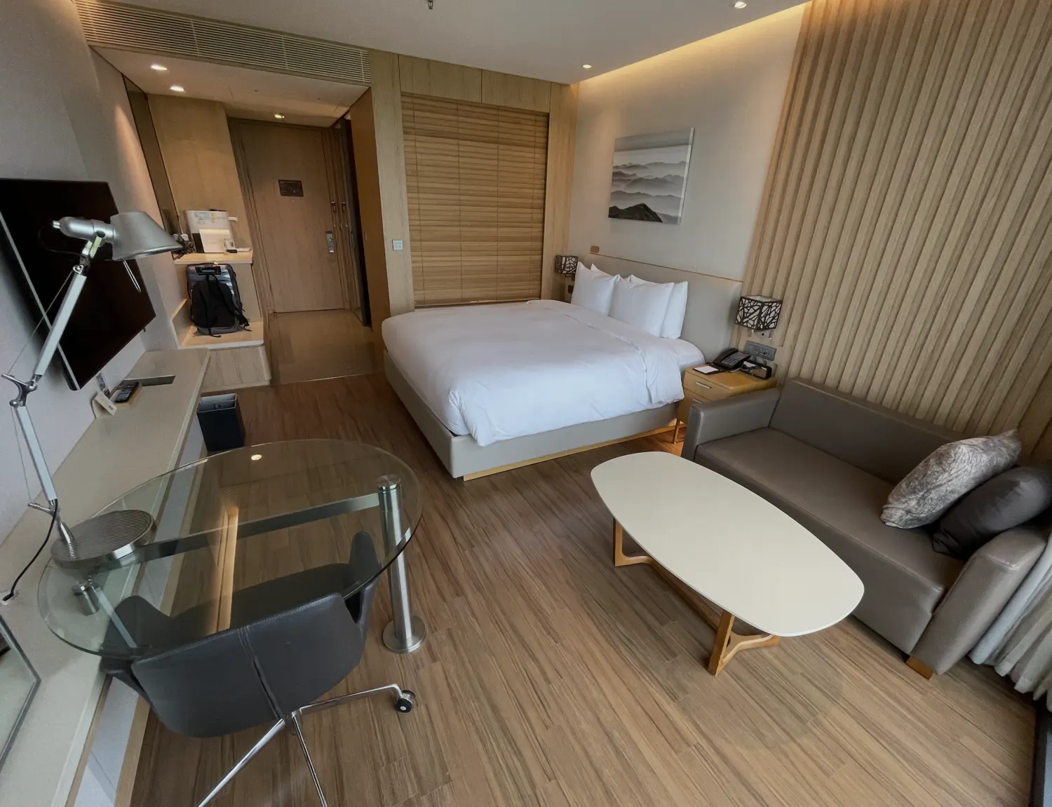 A hotel guest room with a wood-slat wall, warm ceiling cove light, and a blackout shade behind the bed