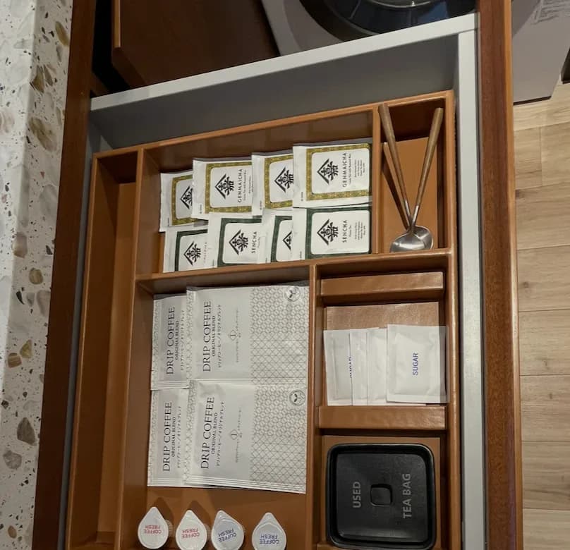 An organized tea drawer with sencha and genmaicha sachets, drip-coffee packets, sugar, and a used-teabag caddy